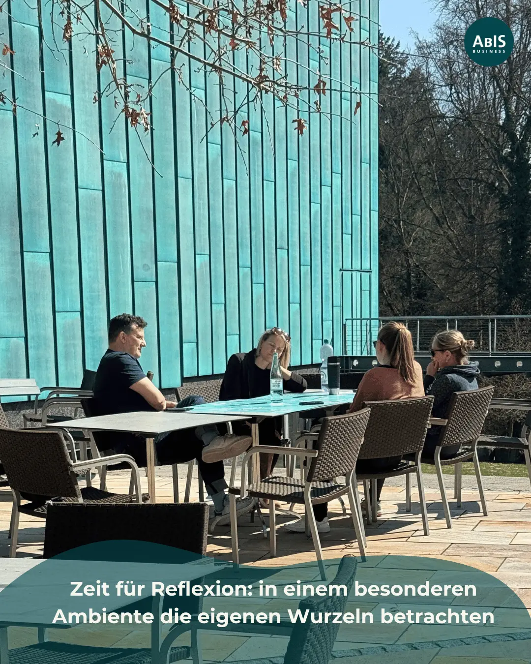 Teilnehmende sitzen draußen an einem Tisch vor einer türkisfarbenen Wand und reflektieren in einer Kleingruppe persönliche Themen.